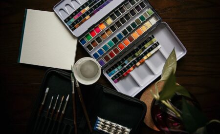 ダークな机の上に置かれた絵筆、水、パレットなどの水彩絵の具セットと植物のある画像